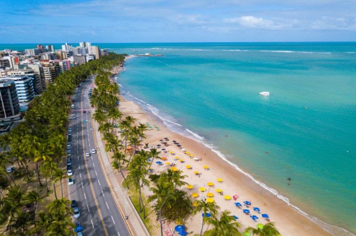 Fotografia aérea da cidade de Maceió na orla, com vista do mar, baaras, a avenida com veículos e edifícios. A água esverdeada e o céu azul
