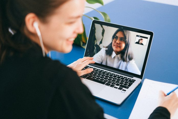 Fotografia de jovem em conferência com uma mulher no notebook. Ela está com fone e escreve em bloco de papel