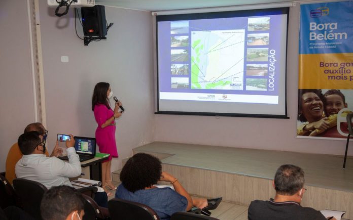 Fotografia da apresentação do projetos estruturantes de mobilidade da cidade de Belém, com uma mulher falando ao microfone fazendo referencia às imagens de slides no telão. Algumas pessoas na plateia acompanham a ação