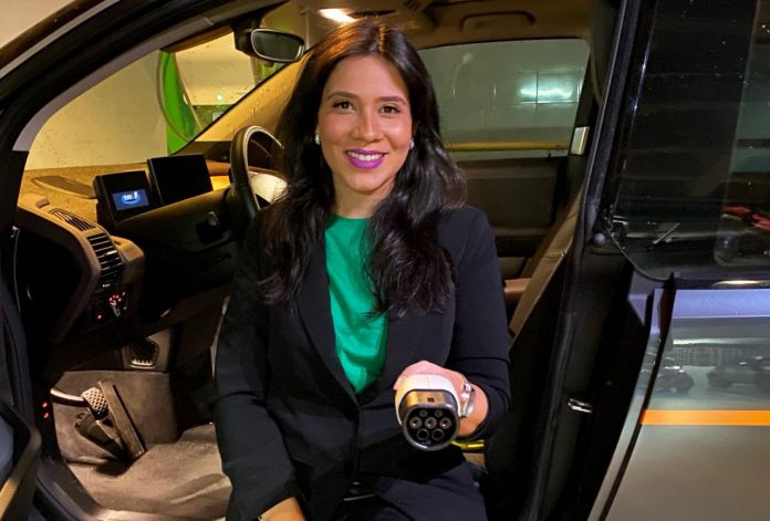 Fotografia de executiva sentada no banco de carro elétrico com as pernas para fora e segurando o carregador do veículo
