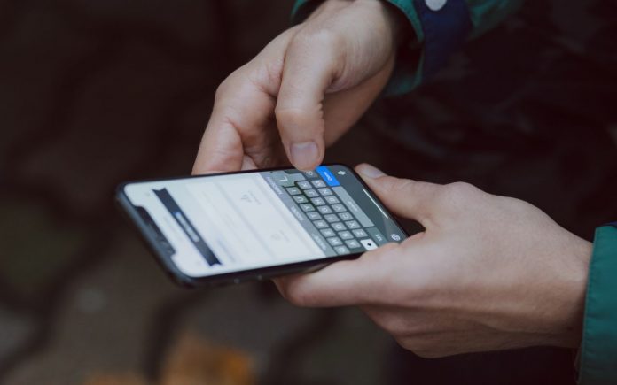 Fotografia com fundo escuro na tonalidade marrom, com pessoa digitando ao celular. Foco nas mãos e no aparelho e levemente nas mangas do casaco verde com detalhe azul