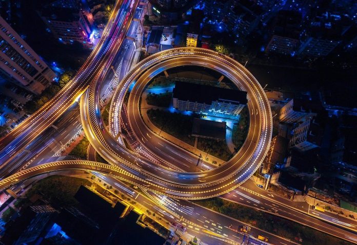 Fotografia aérea com fundo escuro de sistema viário de uma metrópole, onde as várias conexões interligadas são destacadas pelo tom dourado da iluminação pública. Veículos e edifícios também compõem o cenário que contextuliza com cidades inteligentes