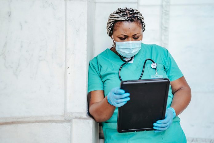 Fotografia de fundo claro com profissional da saúde, com uniforme hospitalar verde, luvas azul e estetoscópio, em cena que remete à pandemia da covid-19. Com expressão de preocupação e usando máscara, ela está lendo e um tablet